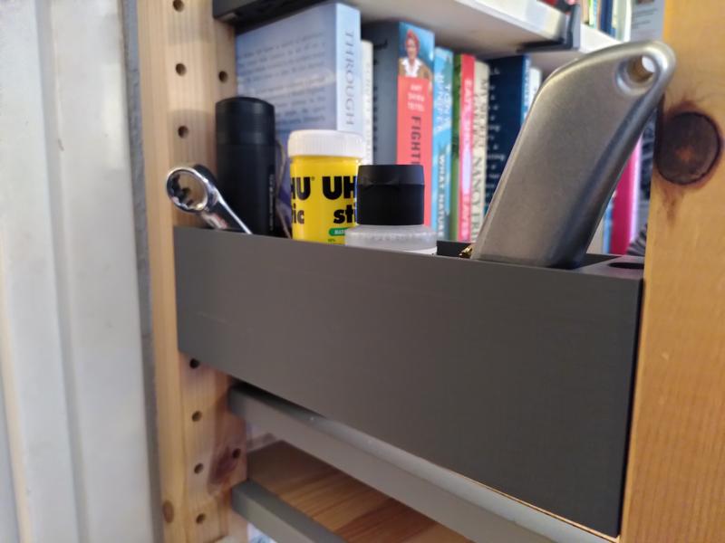 A grey 3d printed plastic bucket between two wooden uprights. Inside the bucket there's the top of a black torch, a ring spanner, some UHU glue, a black plastic cap on a bottle, and a stanley knife. In the background there's various books.