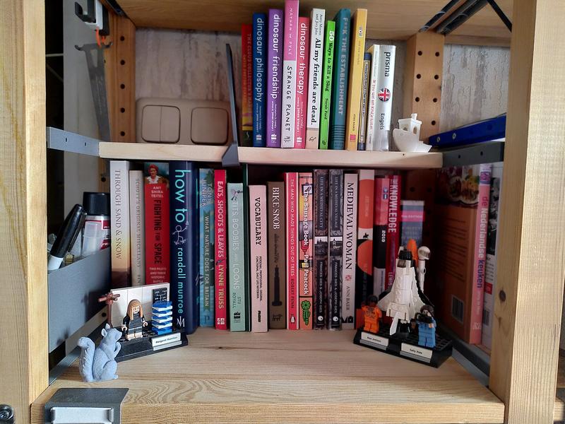 A pair of book shelves, the bottom one is twice the depth as the top one. The top one is held in place with a pair of 3d printed plastic brackets in grey. The top shelf has a number of small books, held in place with a 3d printed bookend, on the other end is a white PLA Benchy. On the lower shelf in front of the shelf are two small lego scultures, one of a space shuttle with a pair of astronauts, and one of Margaret Hamilton next to her source code. There's a 3d printed grey plastic bucket on the left between the shelf uprights. In front of Margaret Hamilton is a small 3d printed grey squirrel.