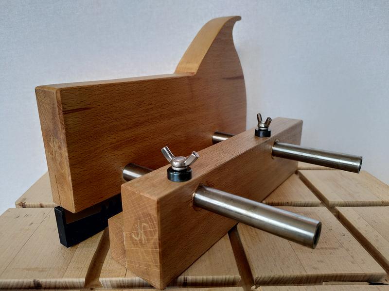 A finished wooden kerfing plane sat on a beech work bench. The kerfing
plane has a dark oiled finish. There is an ambigram JF markers mark
stamped into the end of the fence and main body of the plane. The two
thick washers under the wingnuts on the fence are a dark blue. The main
body of the plane looks like a wooden saw.