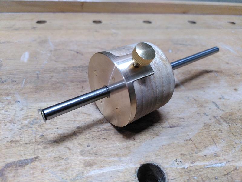 A woodworking marking gauge made of wood (ash - fraxinus excelsior), brass, and polished steel. It sits on a wooden workbench.